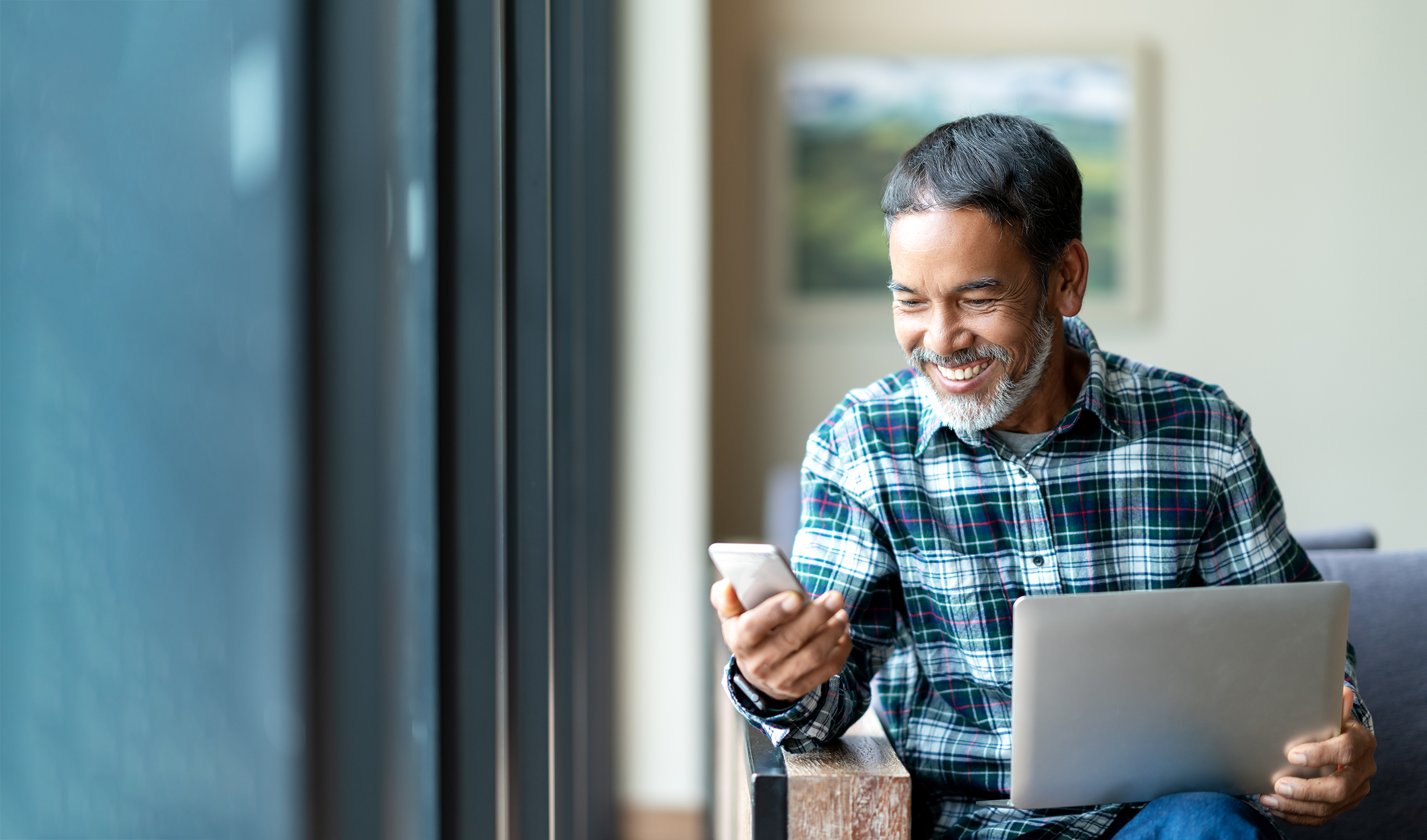 Mature Hispanic adult man smiling with phone and laptop computer