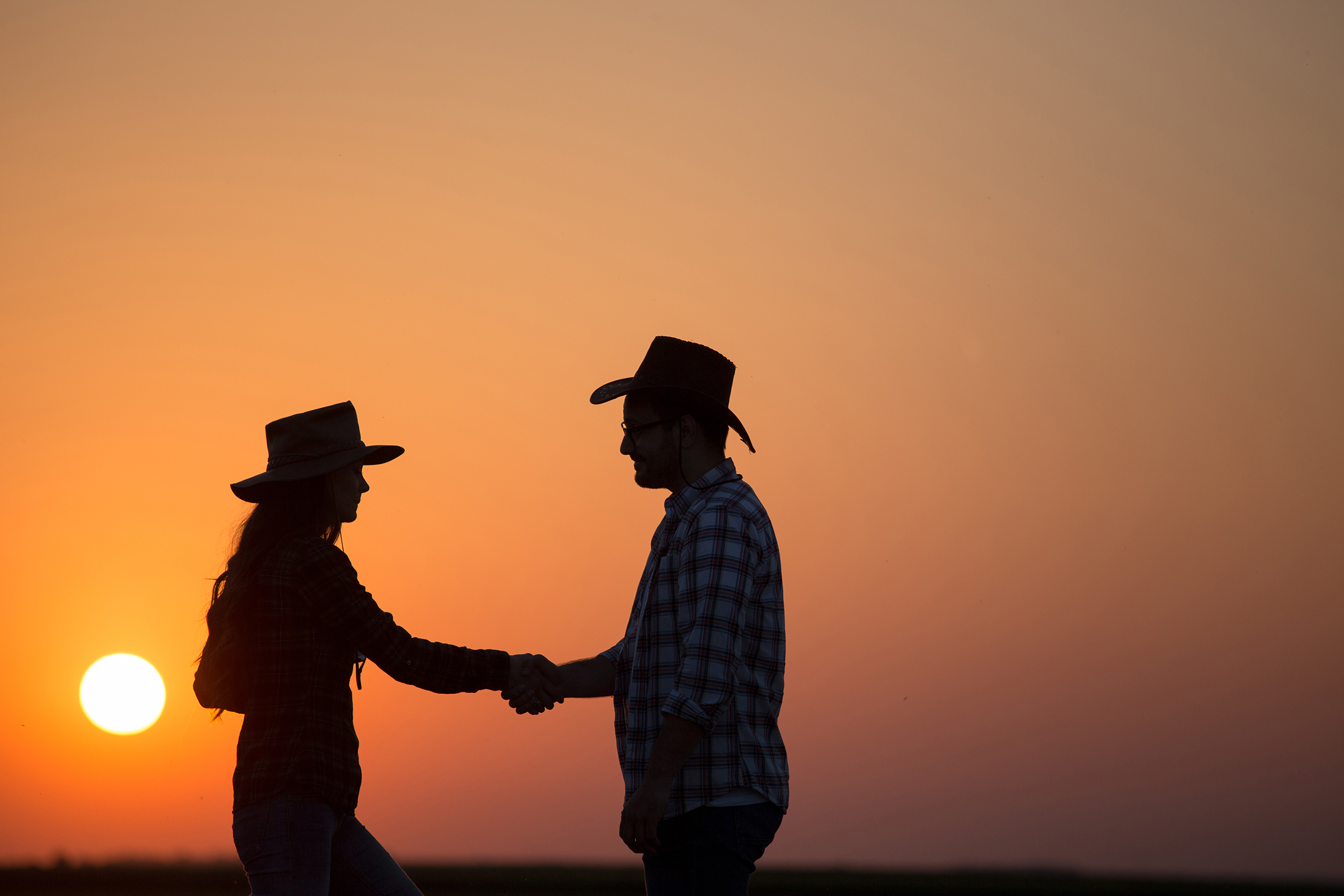 Silhouettes of man and woman farmers with hats shaking hands in field during sunset