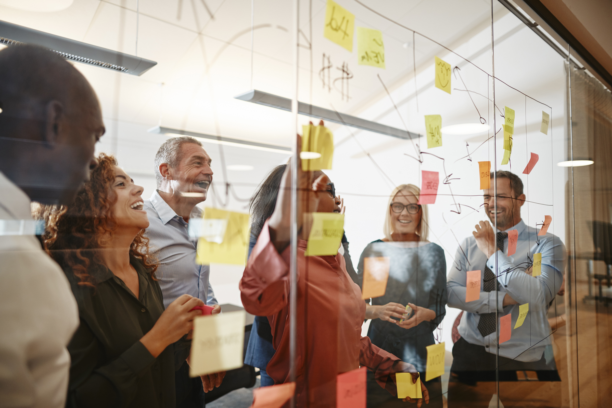 Young African businesswoman and her diverse team laughing while having a brainstorming session with sticky notes on a glass wall in a modern office.