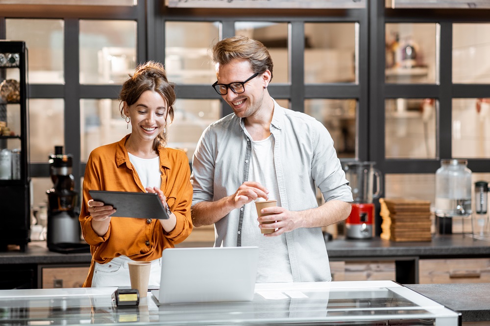 Two young managers or shop owners having some discussion while standing with a digital tablet at the counter of the shop or cafe