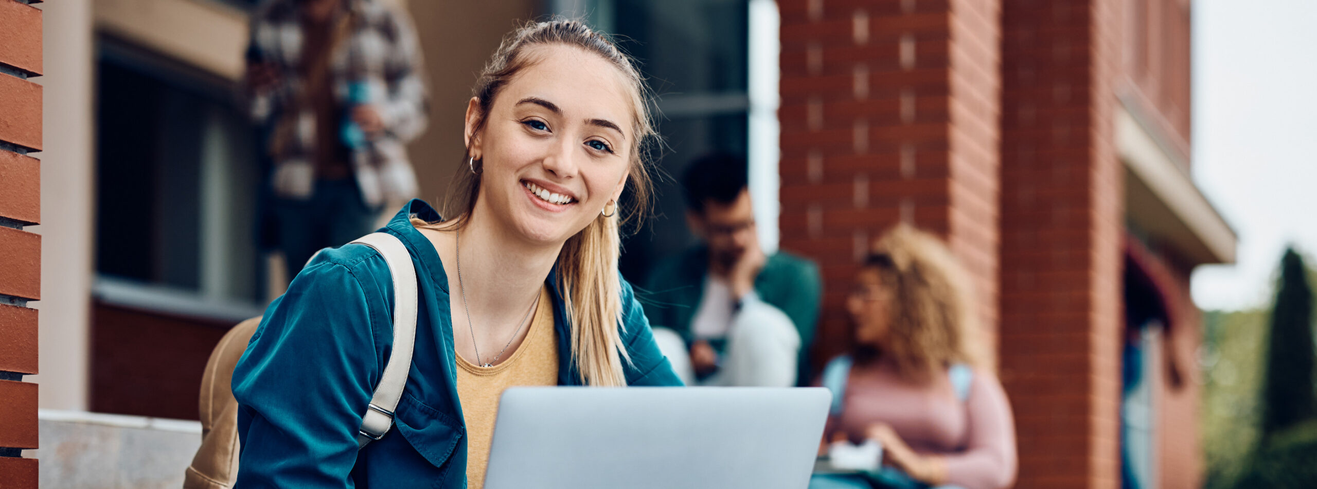 Happy female college student e-learning on laptop at campus and looking at camera.